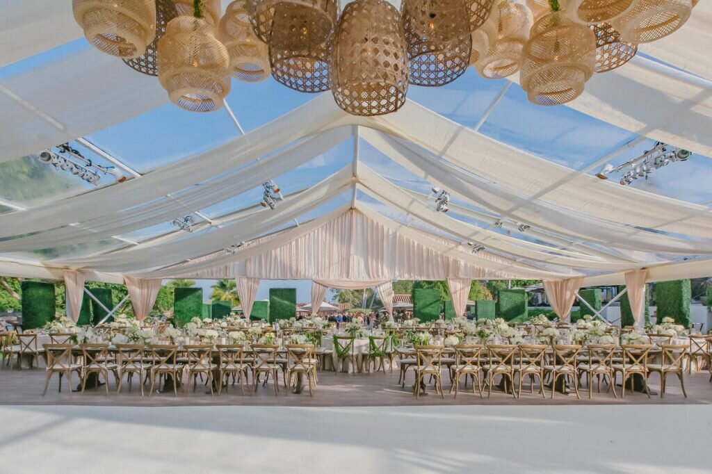 Wedding reception space with a flowing white canopy above, long wooden tables, and wicker chandeliers hanging from above.