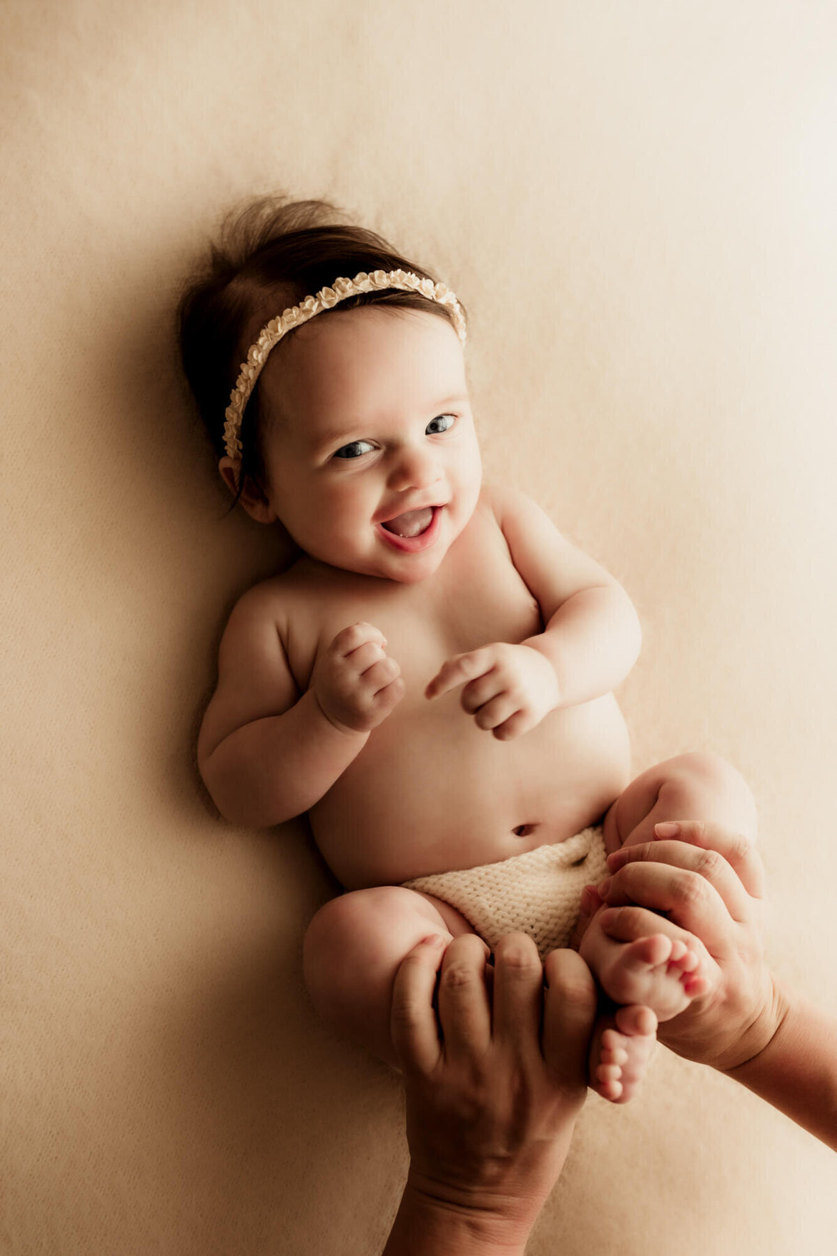 Baby girl wearing a floral headband and laying on her back smiling while her mother holds her legs.