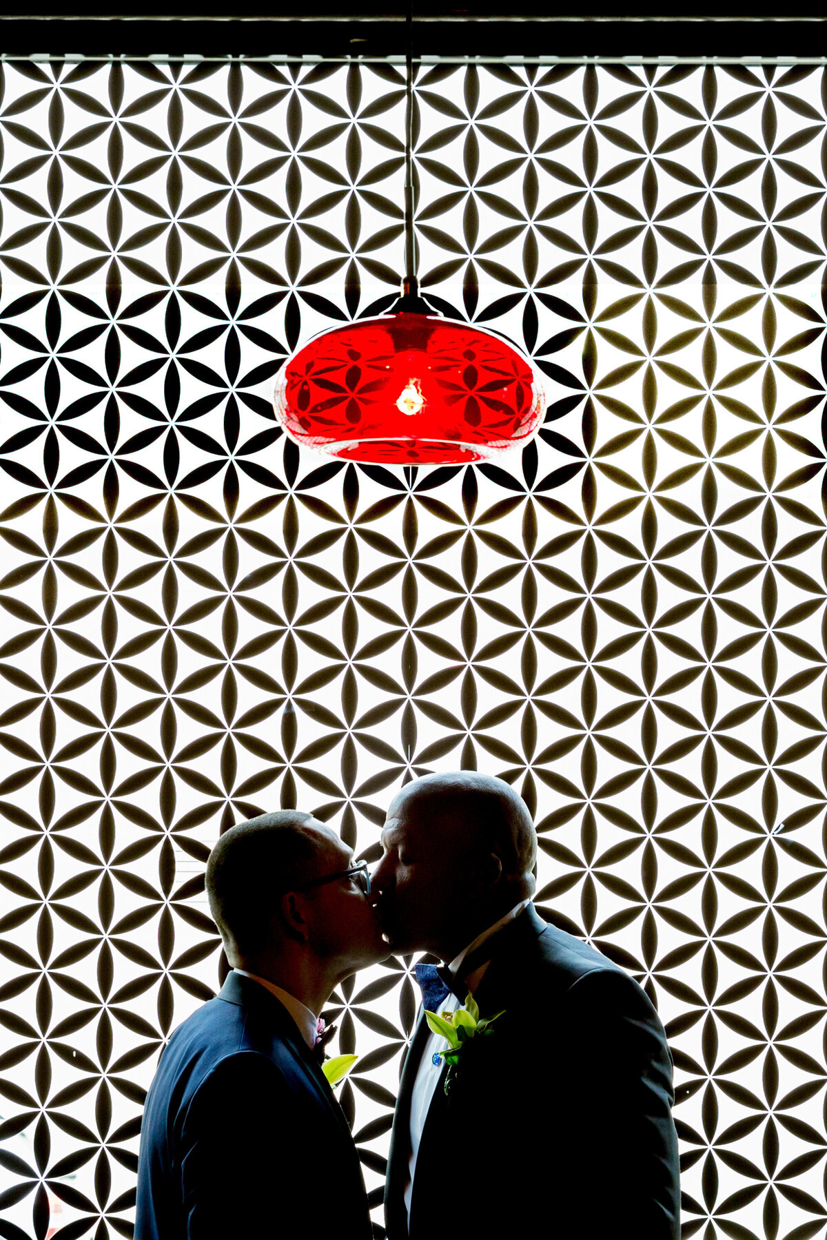 Two grooms kissing in front of a black and white patterned wall.