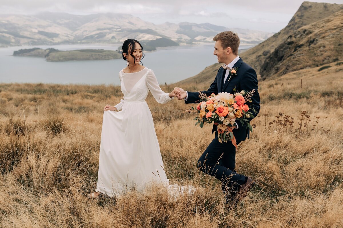 bride and groom walking in long dry grass on top of port hills christchurch with orange bouquet and white dress and navy suit overcast day view of lyttelton harbour