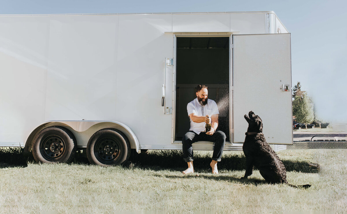 Ryan Erickson celebrating with champagne with his black lab, Henry in front of his mobile portrait studio