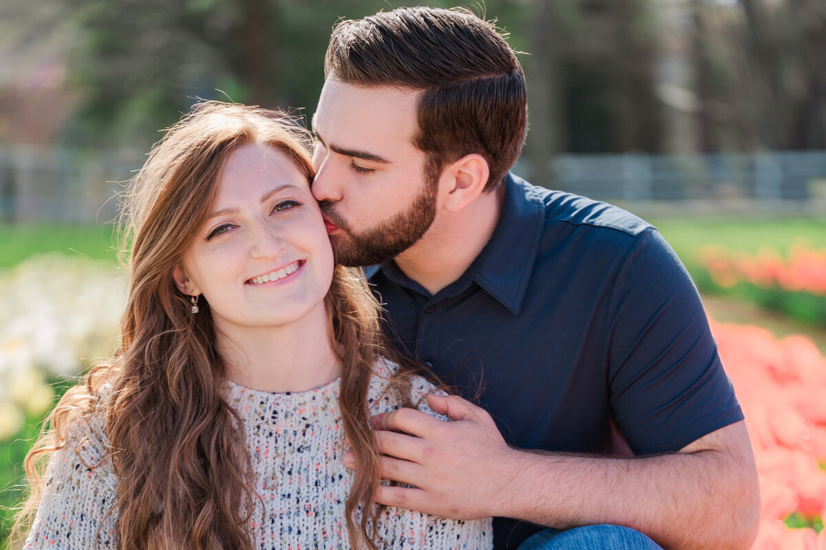 An engaged couple sitting in rows of tulips in the spring sharing an embracing kiss on the cheek by Raleigh wedding photographer, JoLynn Photography