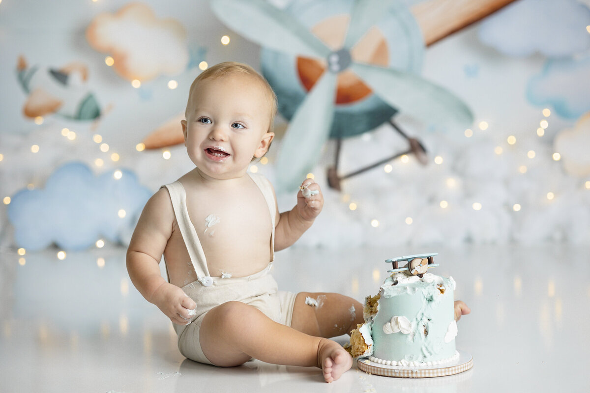 columbus-dayton-ohio-cake-smash-first-birthday-baby-photographer-baby-boy-with-small-airplane-cake-with-vintage-flying-airplane-background-in-light-pastel-blues-amanda-estep-photography