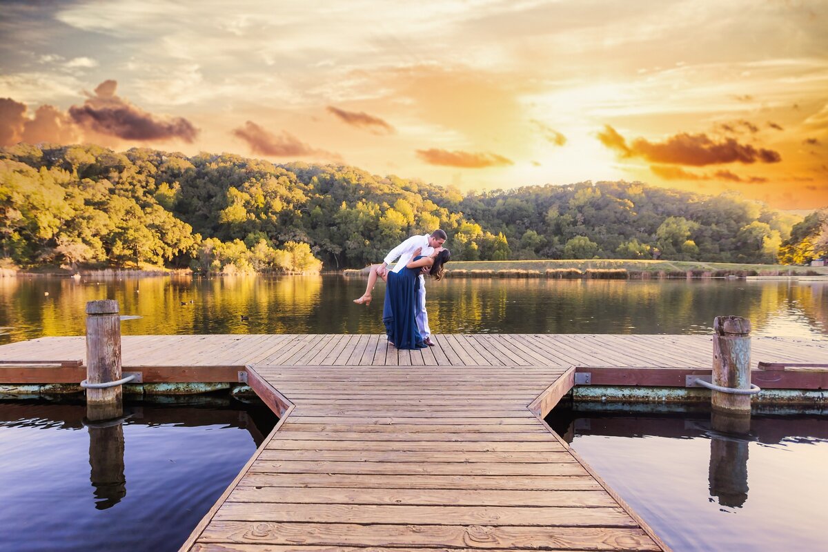 Man dips woman in a blue dress on a dock that sits over a beautiful lake with trees and sunset in the background.  Photo by Sacramento wedding photographer, philippe studio pro.