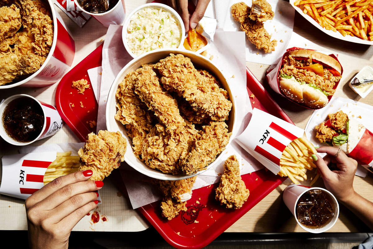 Hands reaching for chicken out of a bucket of fried chicken with fries and chicken sandwiches next to it.