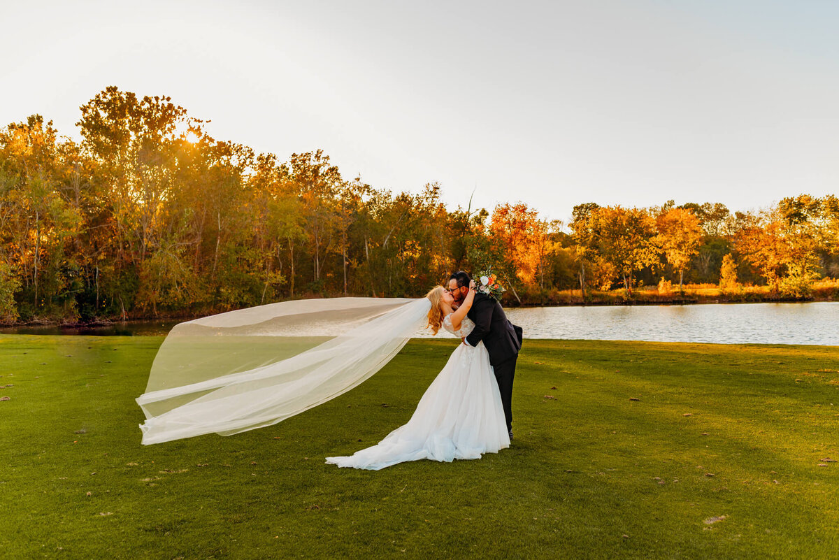 photo of a bride and groom kissing in front of a pond while her veil flows in the wind
