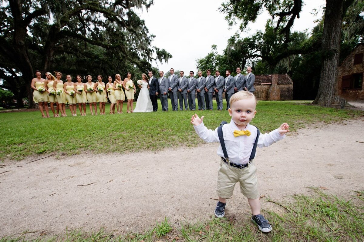 a tiny ring bearer walks toward the camera while the rest of the wedding party are lined up in the background