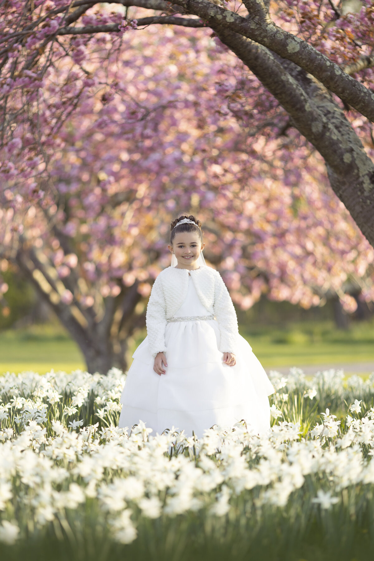 A young girl standsing in a field of white wildflowers and pink blooming trees in a white communion dress
