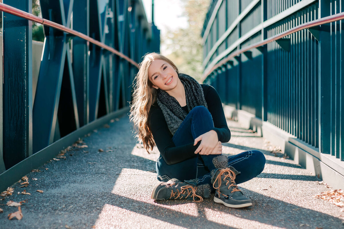 a long blonde haired girl sitting with one leg tucked in and the other pulled to chest on a bridge