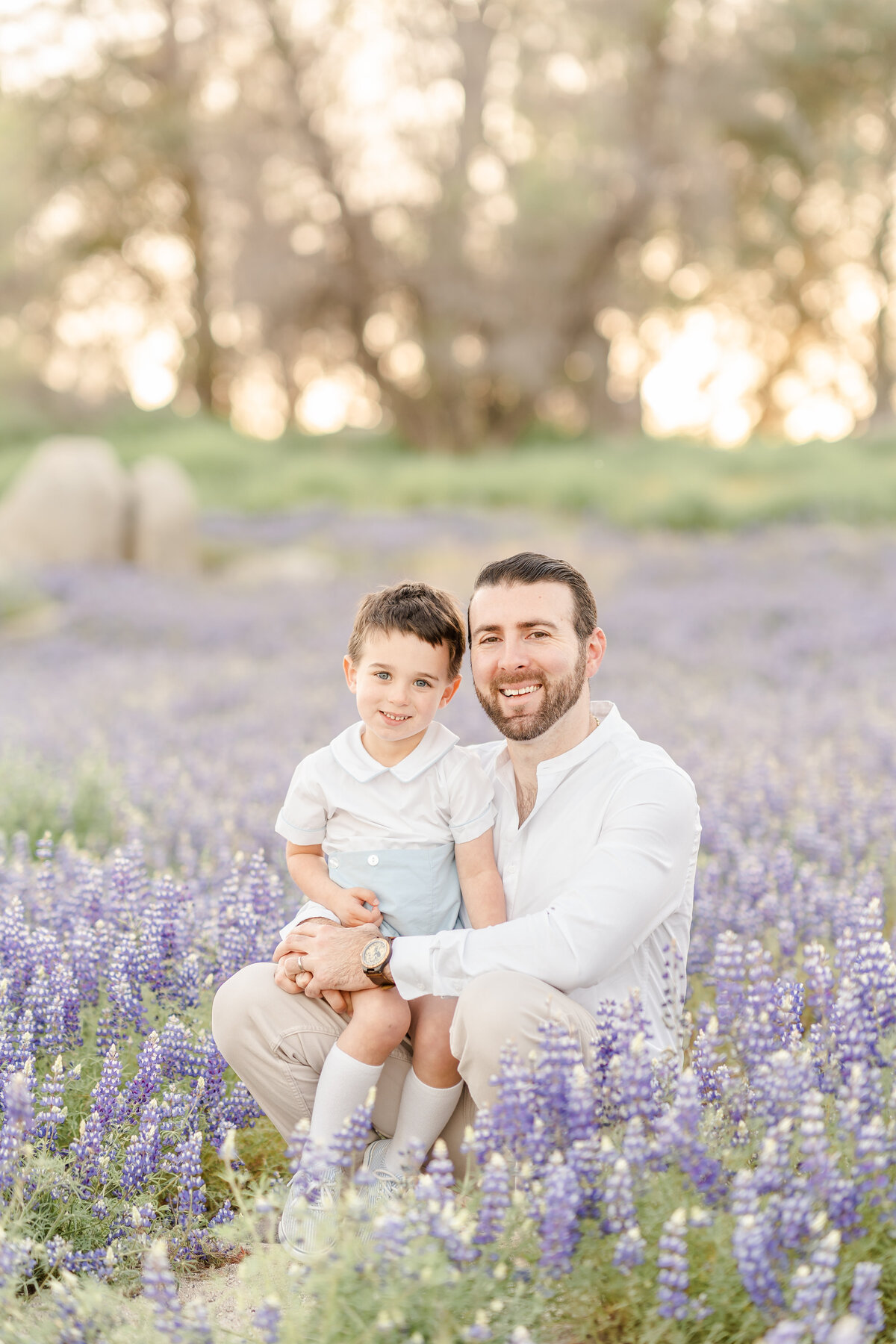 A dad kneels with his son on his lap in a field of lupines