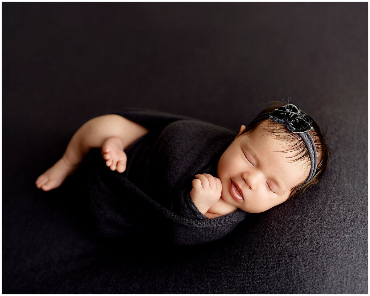A newborn baby laying on a plush gray backdrop and wrapped also in gray wrap and smiling.