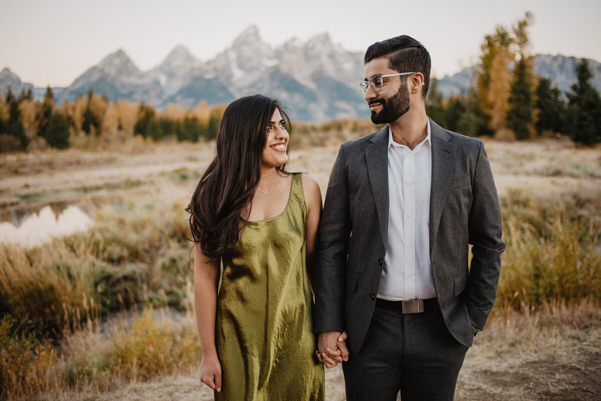 photographers in jackson hole photographs woman in an olive green velvet dress holding hands with a man in a suit and glasses while walking in Jackson Hole for their fall engagement session