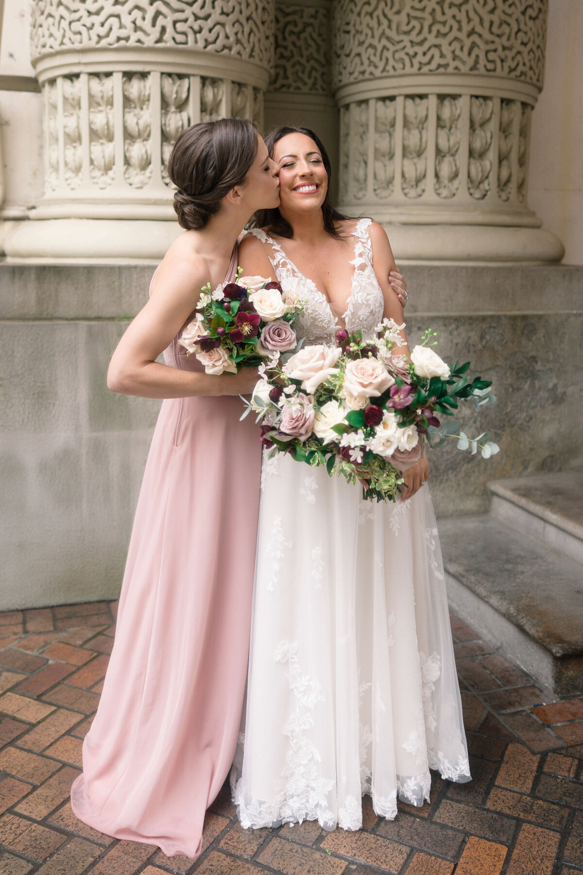 A bride in a white lace wedding dress holds a bouquet of roses while smiling. Next to her, a woman in a pink dress kisses her cheek. They stand in front of ornate stone columns on a brick path.