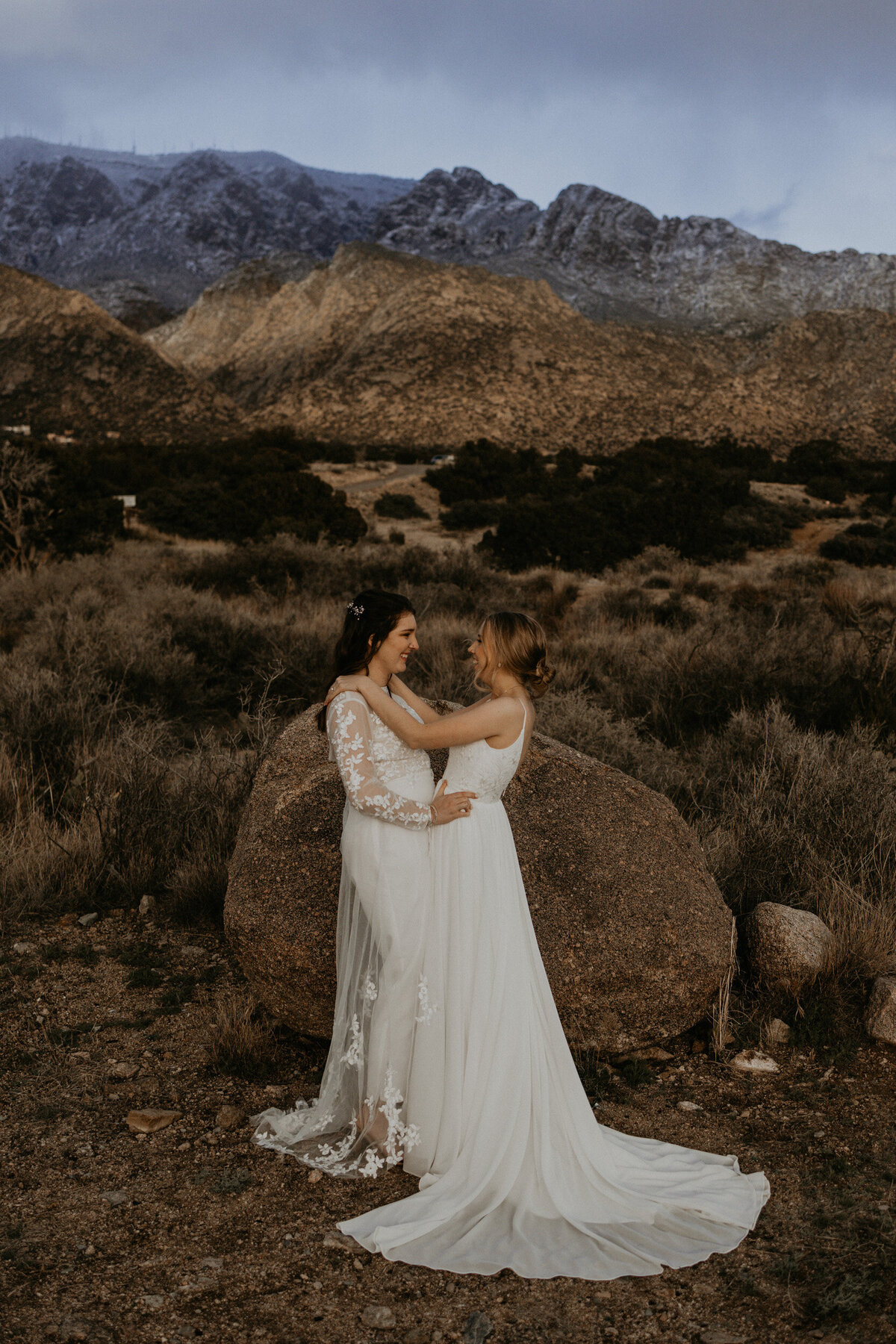 two brides standing together holding each other in the desert