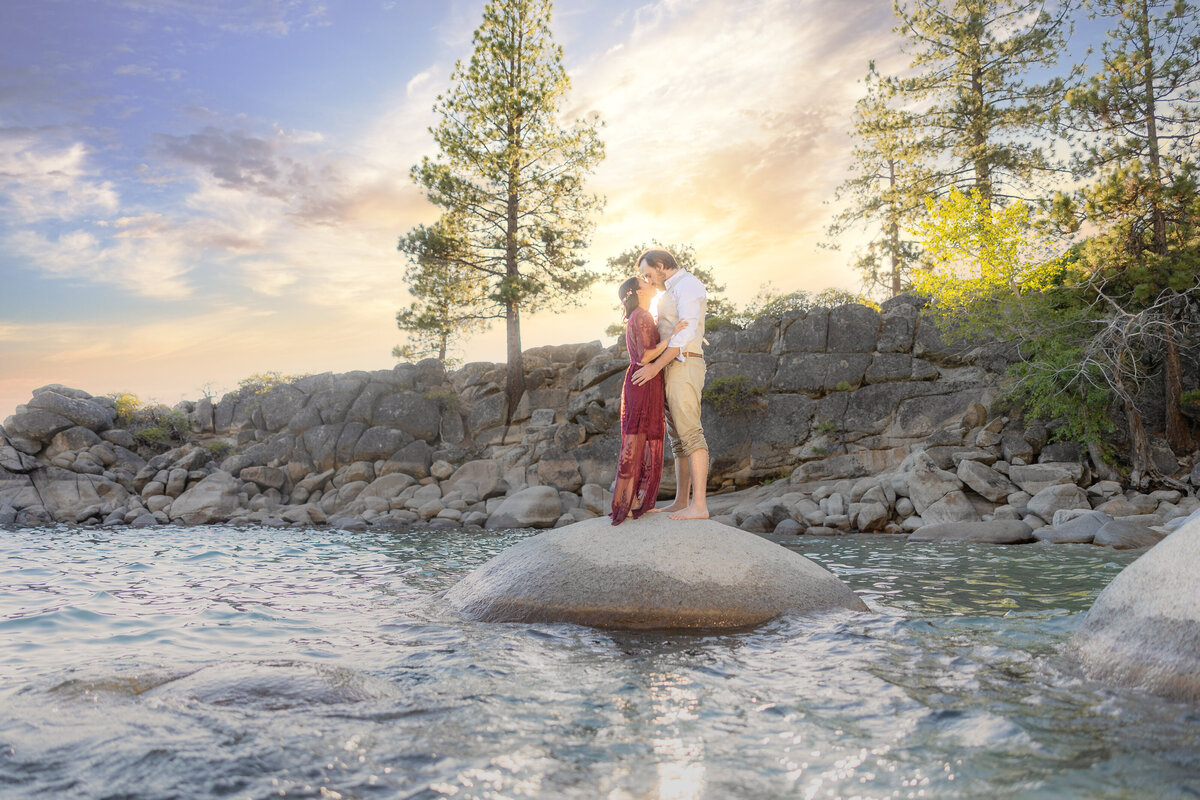 Engaged couple kiss while standing on top of a rock in Lake Tahoe Secret Cove with the sunset in the background. Captured by wedding photographer, philippe studio pro.