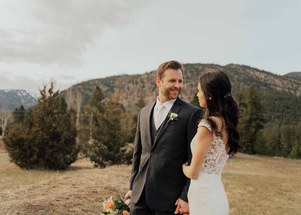 Maddie Rae Photography bride and groom standing side by side. they are looking at each other. he is holding her flowers. there are mountains in the background