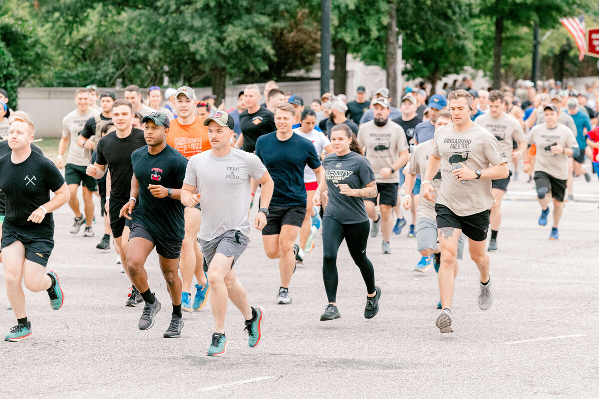 A large group of people is running on a street in a marathon, captured beautifully by an event photographer in Fayetteville, NC. They wear casual sports clothing, and some have caps. The backdrop features trees and a sidewalk, adding to the energetic and lively atmosphere.