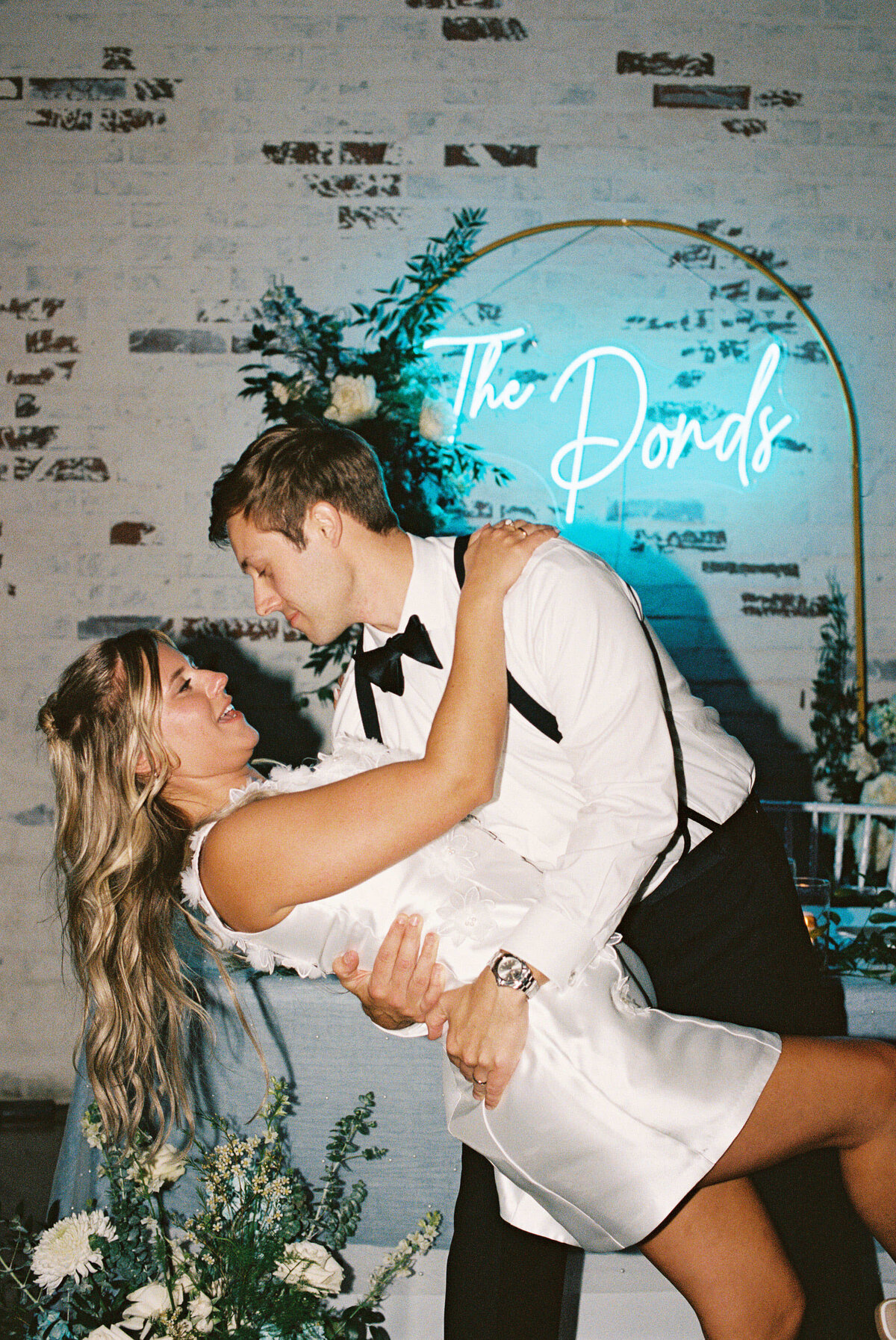 A joyful couple in formal attire shares a dance, the man dips the woman under a sign reading "The Donds," with elegant floral decor behind them.