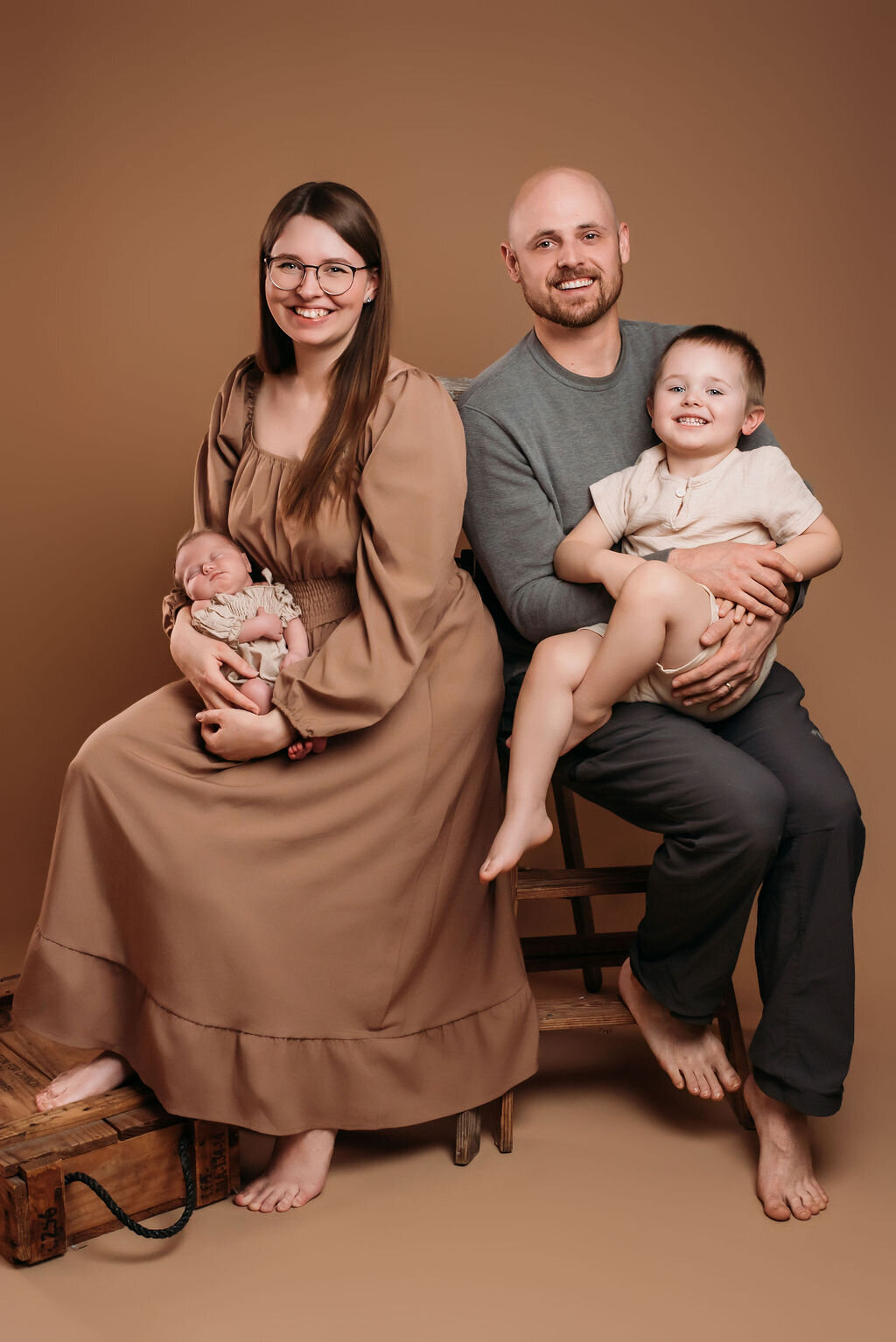 A family sits together barefoot in front of a neutral-toned backdrop—a woman holding a baby and a man with a young child, captured by a photographer in Bemidji, Minnesota.