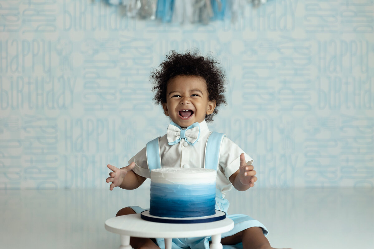 A happy toddler boy in blue suspenders and bowtie sits behind a blue cake for his first birthday