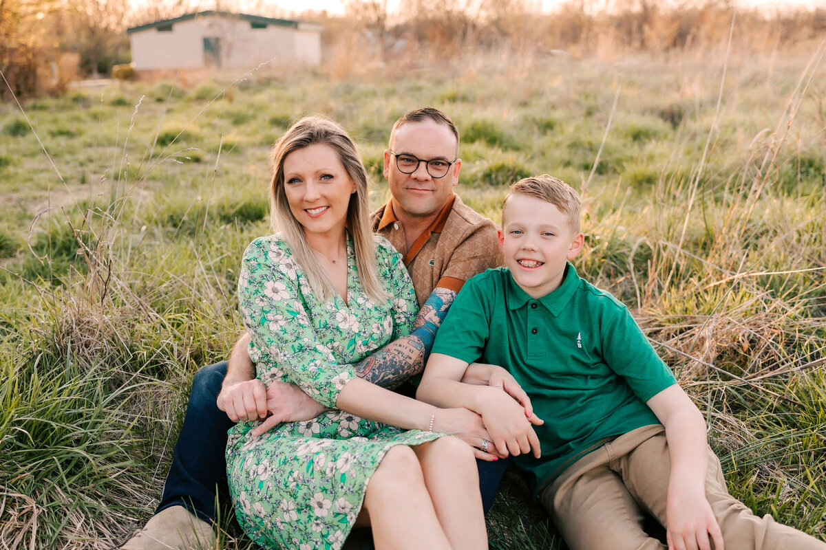 Mom, dad, and son are sitting in the tall grass in the middle of a field at sunset they are looking up at the camera and smiling