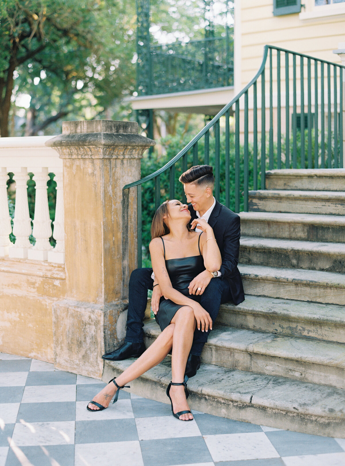 Inclusive wedding photographer based in Charleston, SC. Couple sitting on steps laughing at each other.  Chic and stylish black outfits at sunset engagement session. Charleston film photographer.