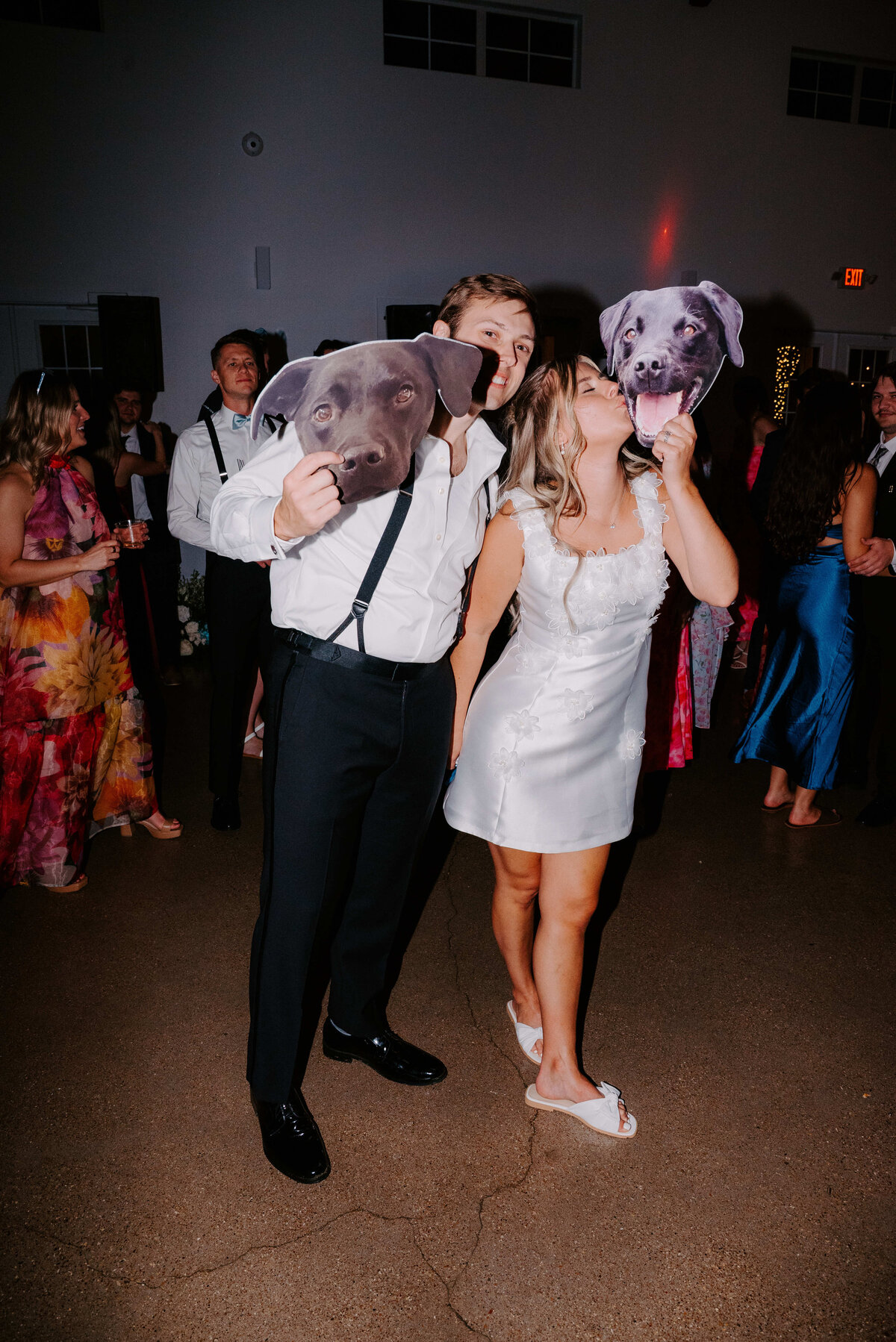 A couple at a party, both wearing formal attire, hold large dog face cutouts in front of theirs, adding a playful, joyful touch to the lively atmosphere.
