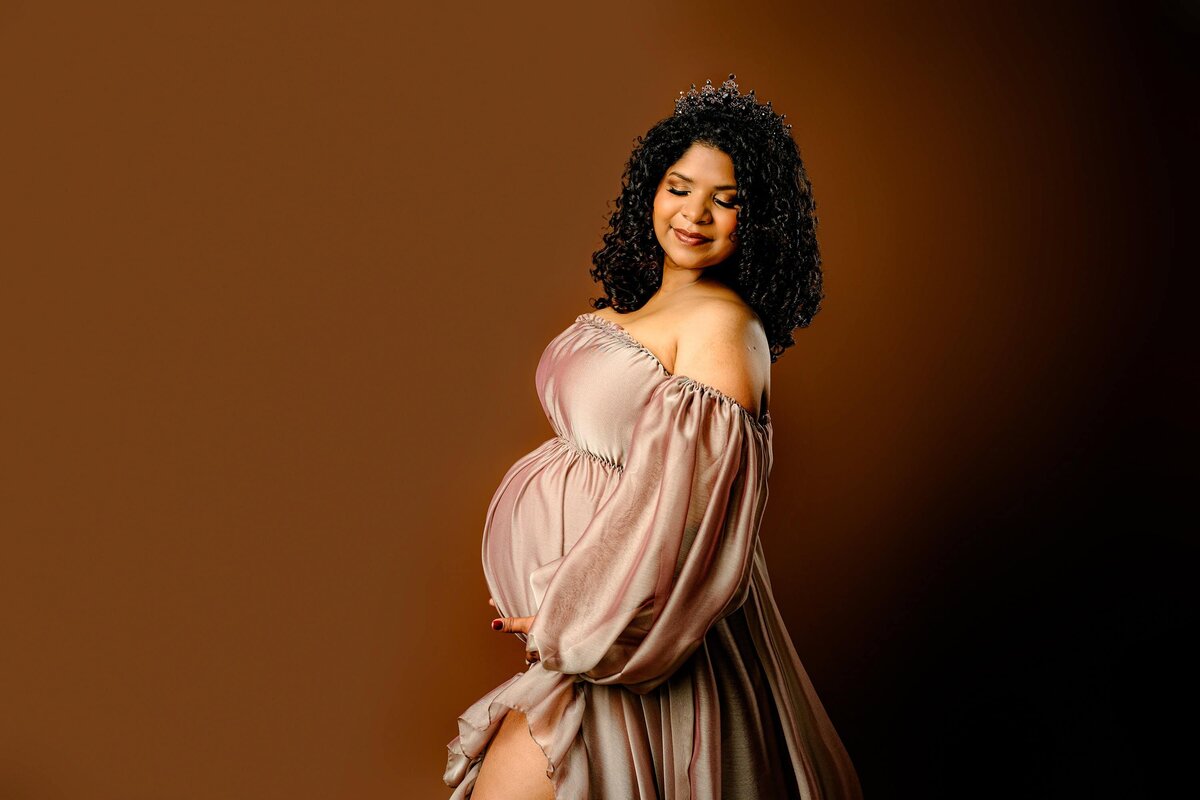 expectant mom dressed in a gold toned off the shoulder  maternity dress on a brown studio background