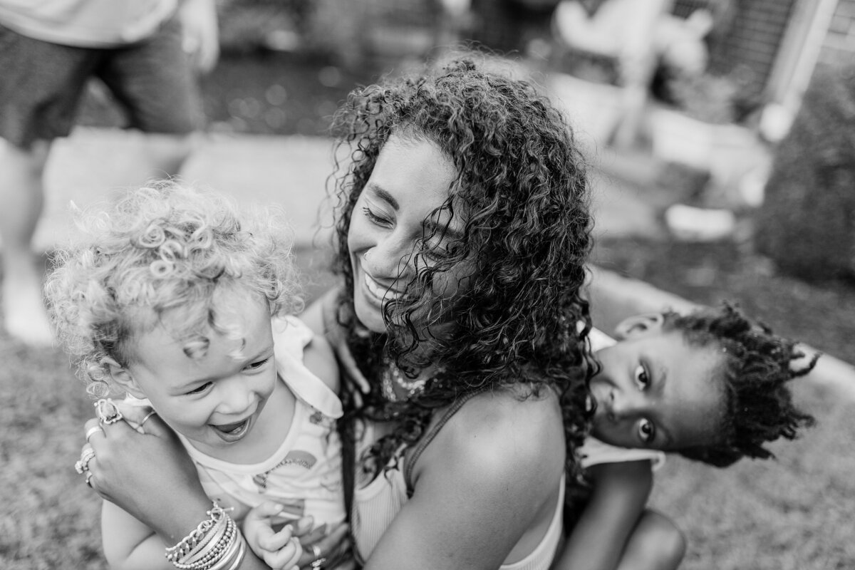 A black and white image of a mom with her two daughters holding onto her during a lifestyle photo session in Lexington KY.