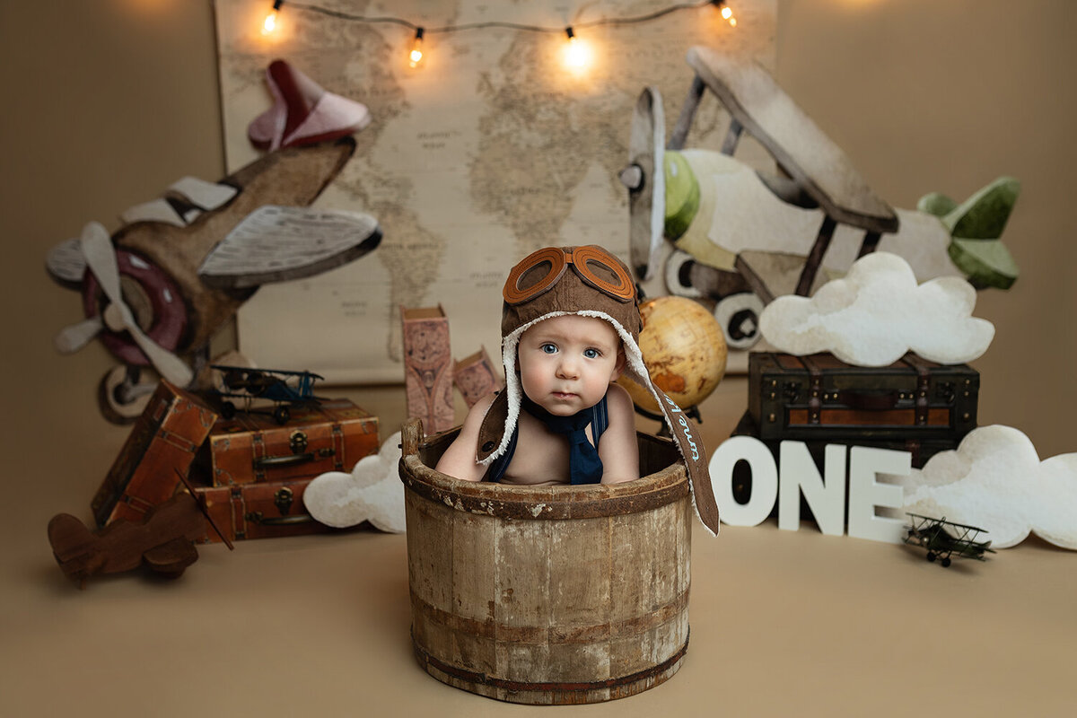 A toddler boy dressed as an aviator sits in a wooden bucket in a themed studio setup