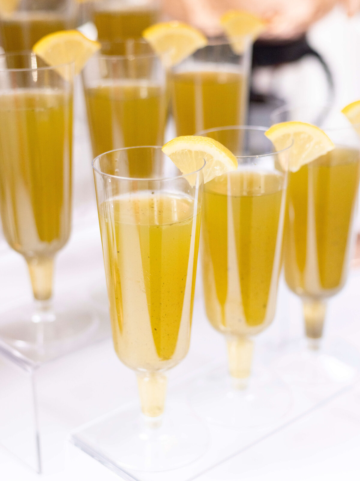 Several tall, clear glasses filled with a light yellow beverage, each garnished with a lemon wedge on the rim, arranged on a white surface.