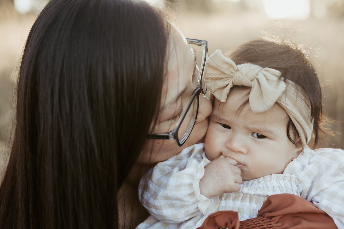 Baby girl wearing a cute headband has her fingers in her mouth and is looking at the camera while her mum is kissing the side of her face.