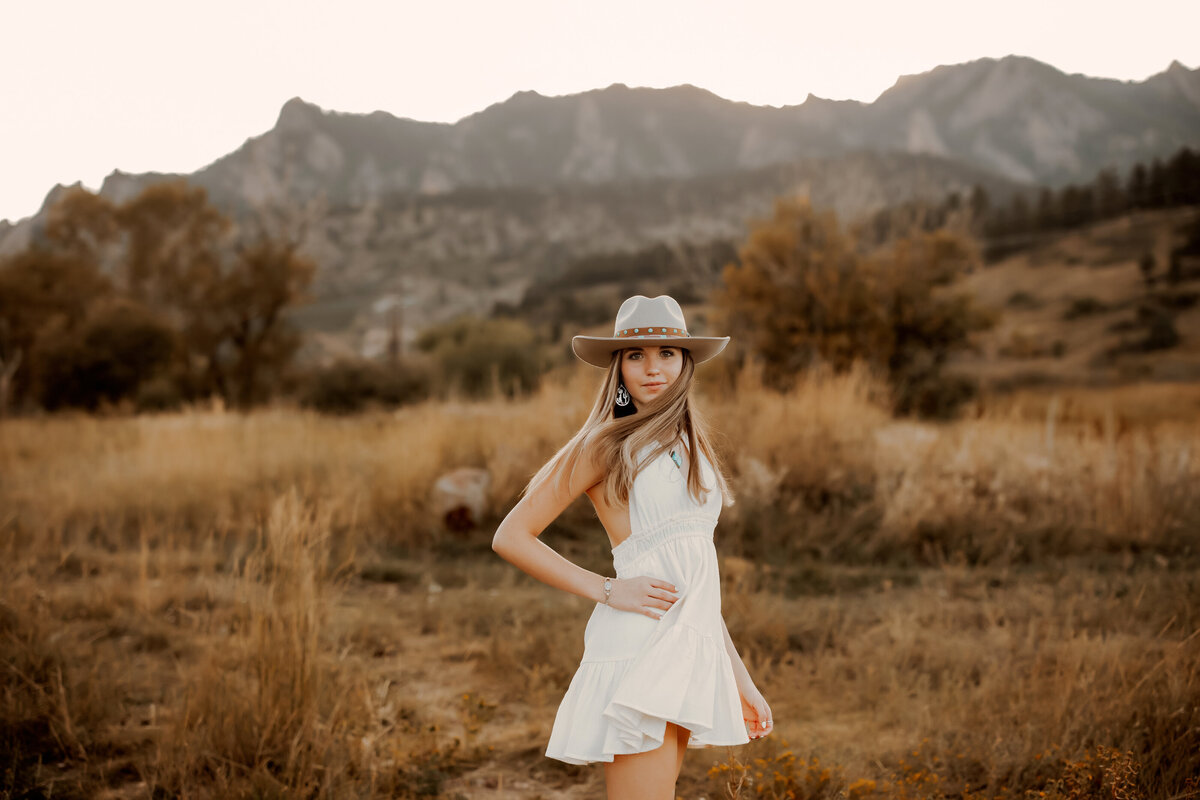 high school senior girl spins in dress and cowgirl hat in boulder colorado with mountains in the background at golden hour