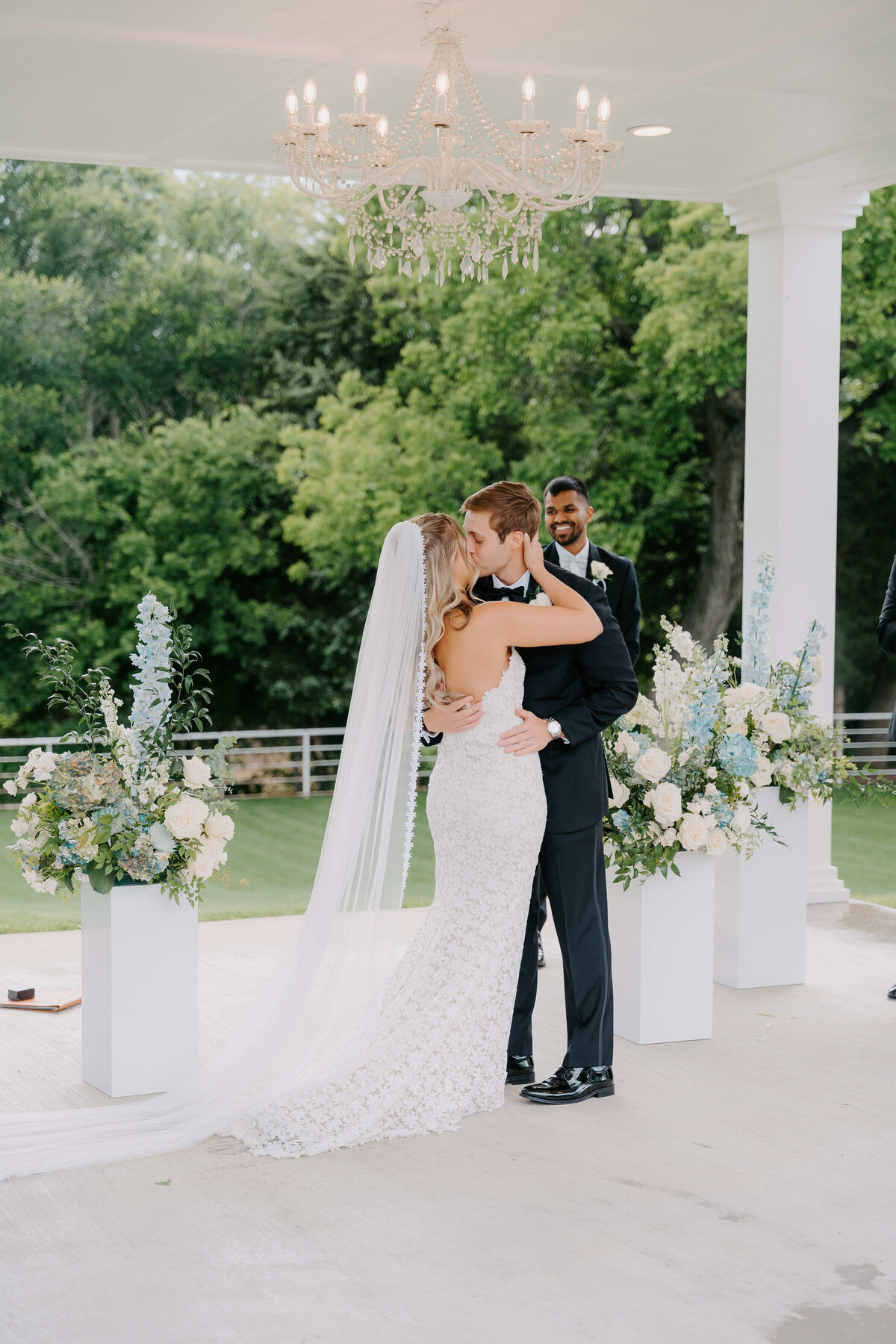 A bride and groom share a kiss at an outdoor wedding, surrounded by lush greenery. Floral arrangements and a chandelier add elegance to the joyous scene.