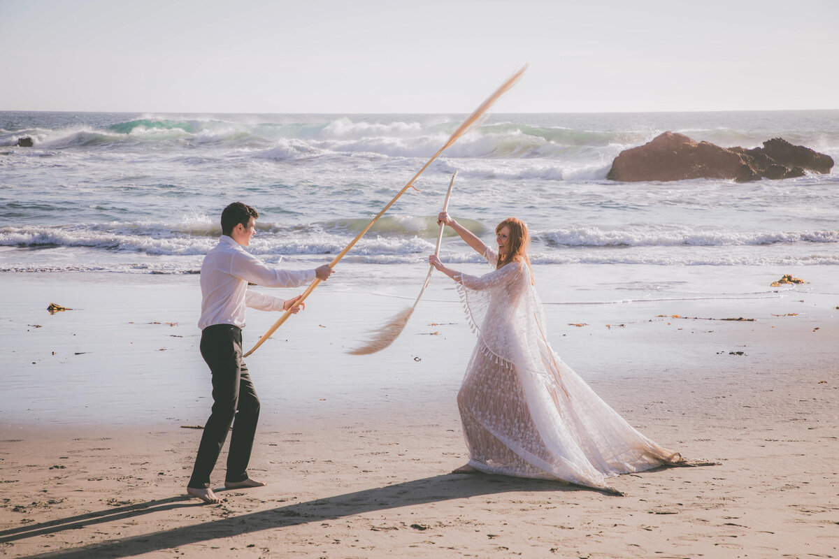 Bride & groom play fights with long cattails on beach.