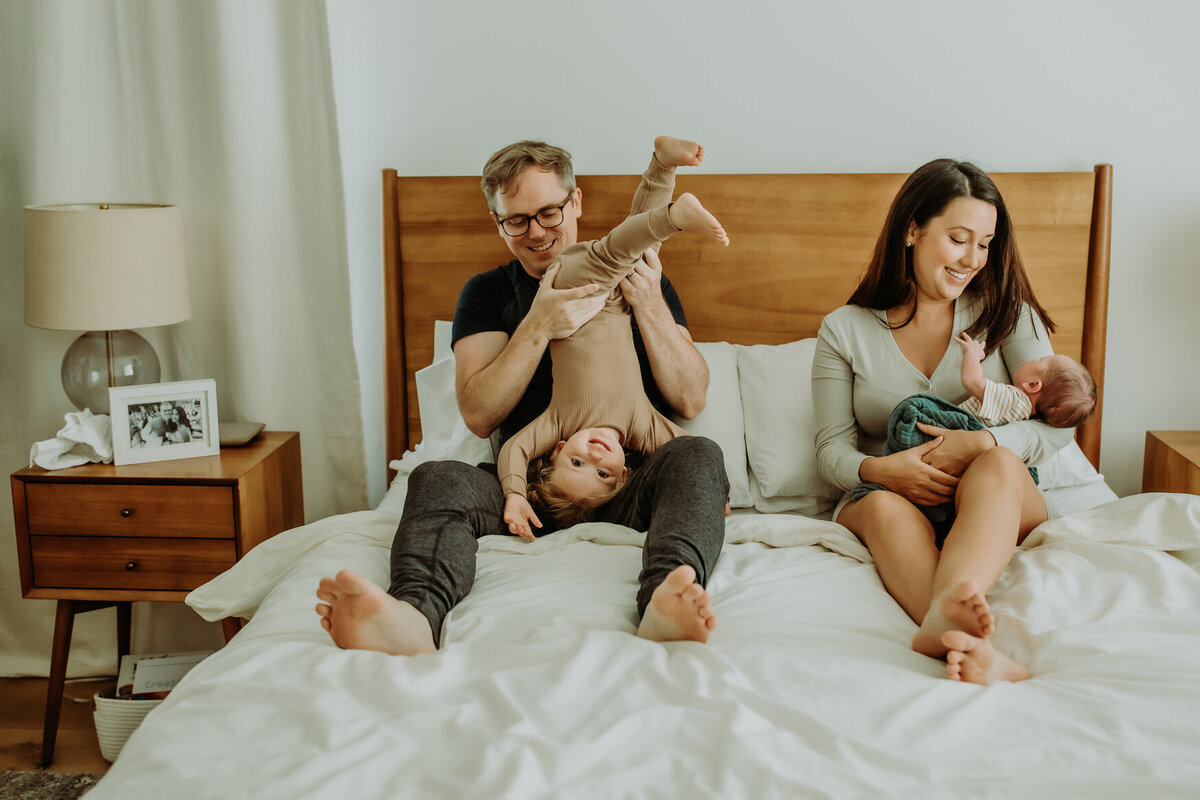 San Francisco family photo with newborn in home on bed with toddler playfully hanging upside down by dad while mom holds baby