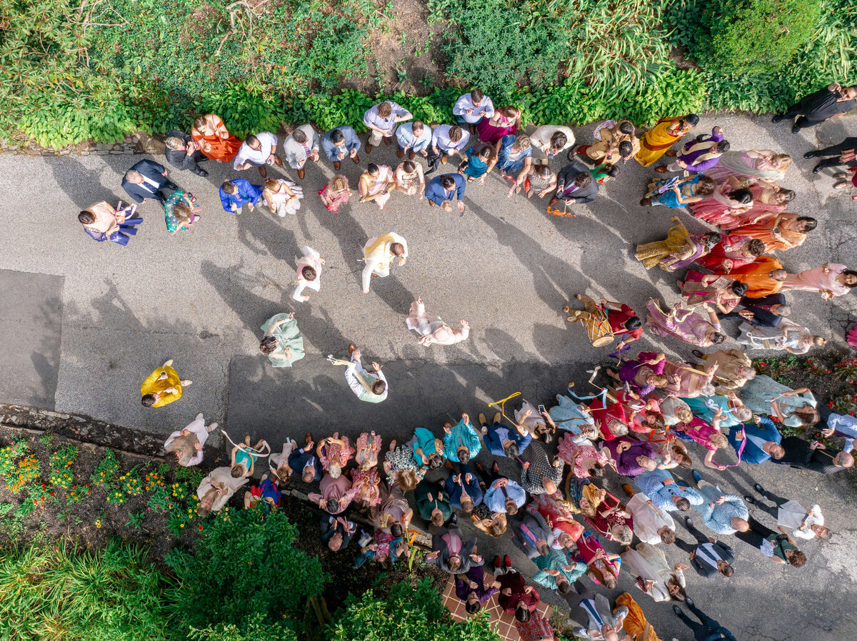 Aerial view of a colorful parade or procession on a pathway, with people wearing vibrant traditional clothing. The gathering takes place outdoors, surrounded by greenery, as participants form a semi-circle, creating an engaging and festive atmosphere.