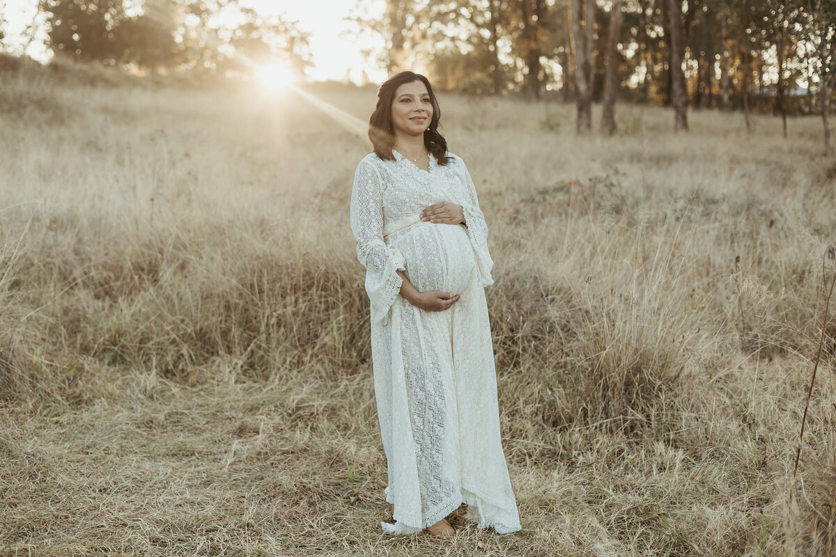 Pretty woman cradling her baby bump while wearing a beautiful lace gown in a gorgeous grassy field while the sun sets behind her