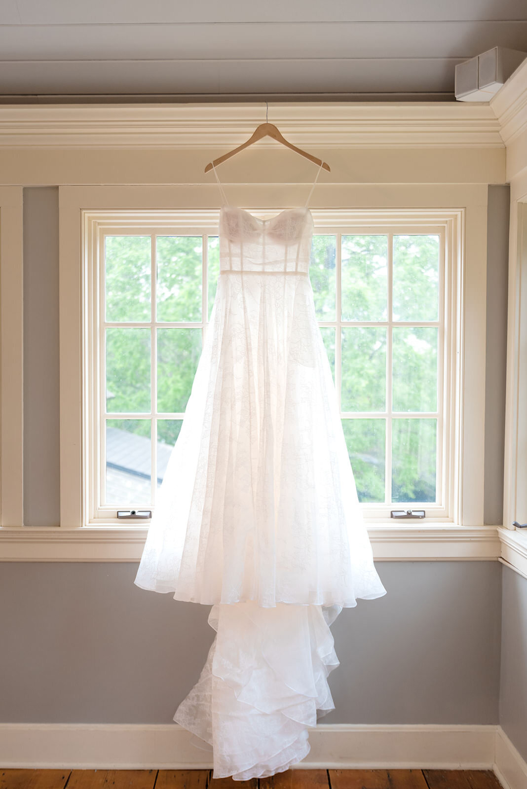 A white wedding dress hanging in front of a window in a room with cream walls.