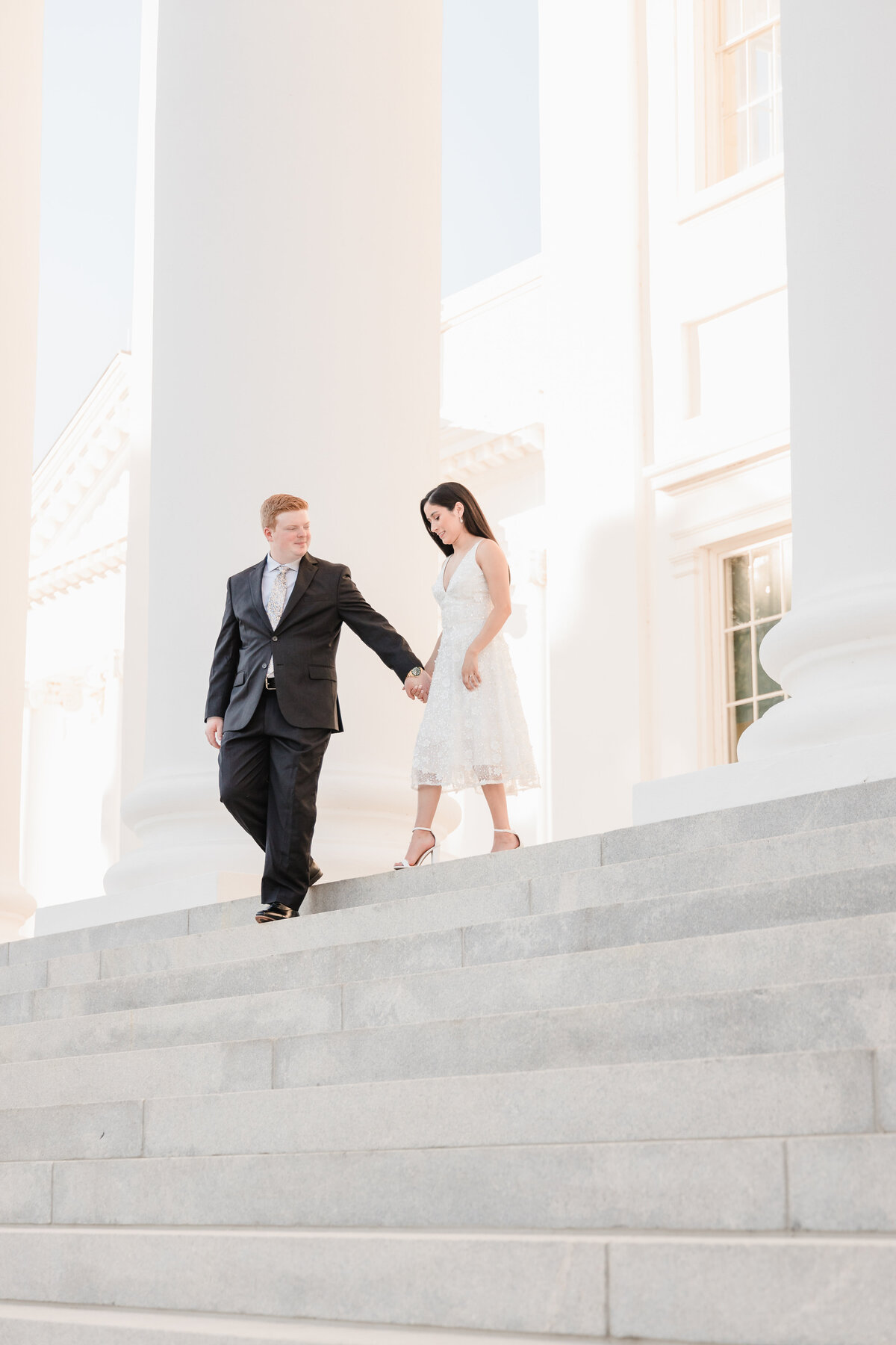 bride in white dress being led down light colored stairs of all white virginia capitol building