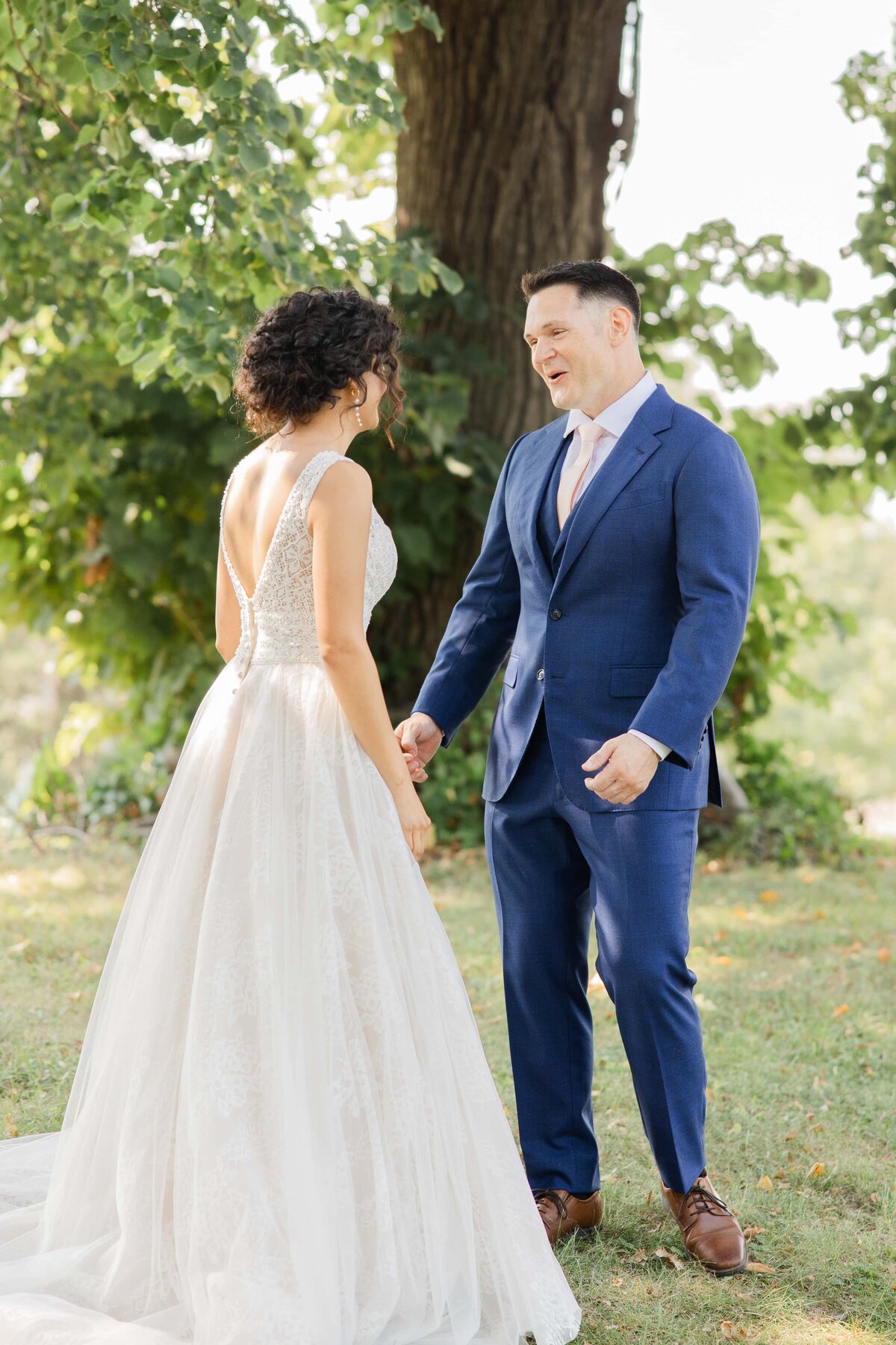 A bride in a white lace dress and a groom in a blue suit smiling and holding hands outdoors near a tree at Park Farm Winery.