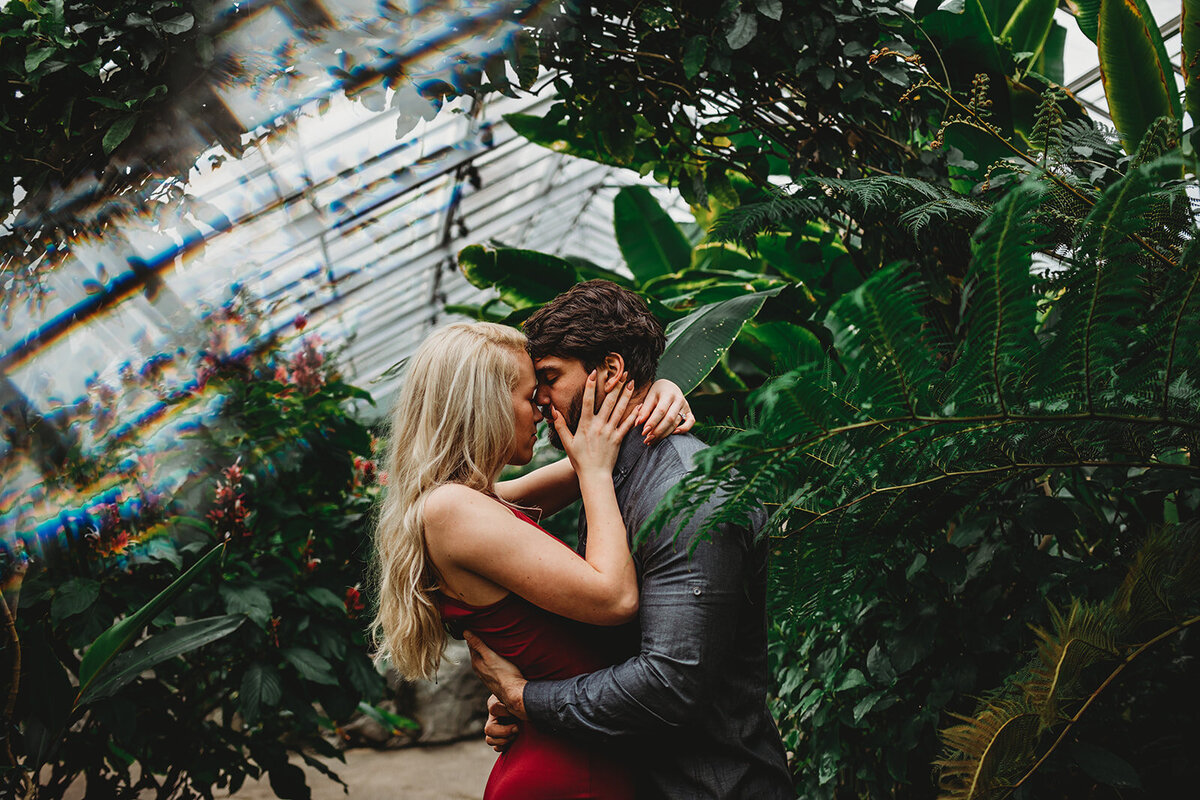 Baltimore wedding photographers captures greenhouse engagement photos with man embracing his fiancé around the waist well she holds the back of his neck romantically