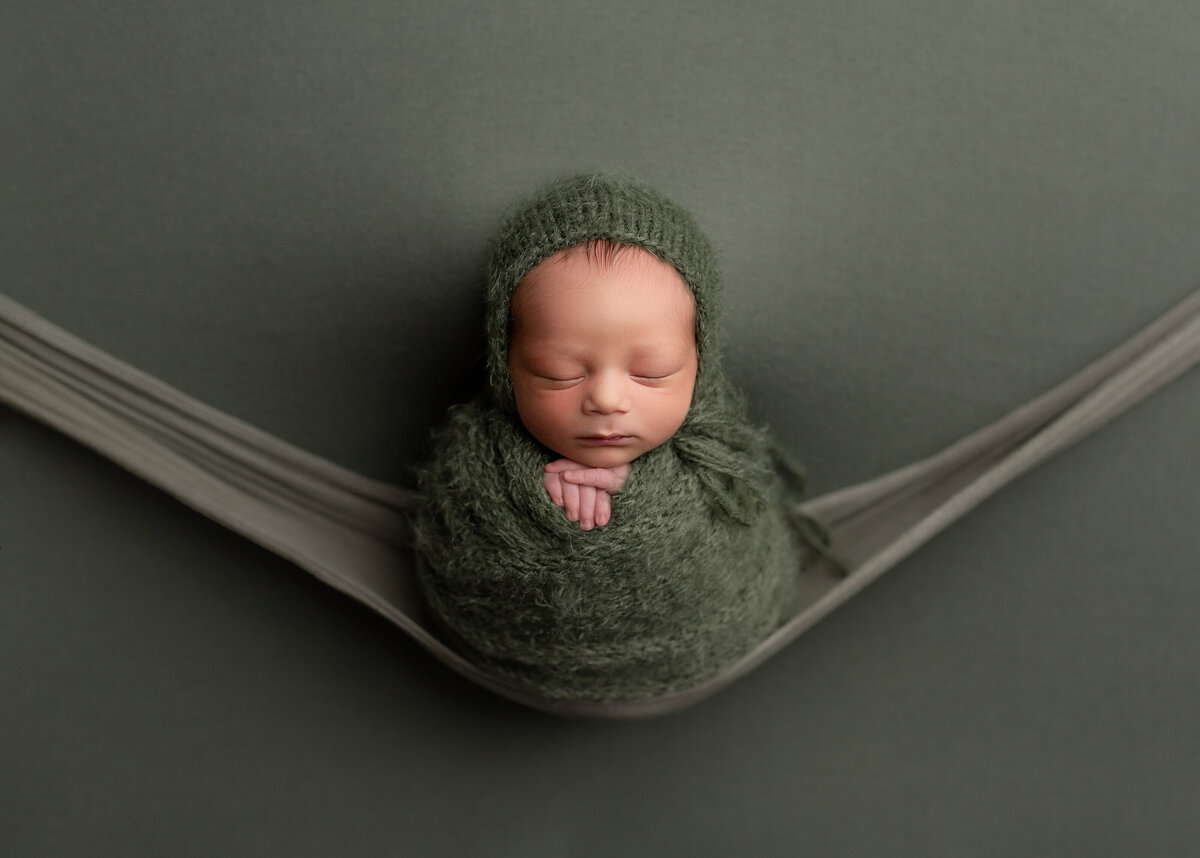 Newborn photoshoot with top West Palm Beach photographer. Baby is wrapped in a green knit swaddle and matching bonnet. Baby is resting atop of a green stretchy wrap, giving the illusion that the baby is resting on a fabric swing.