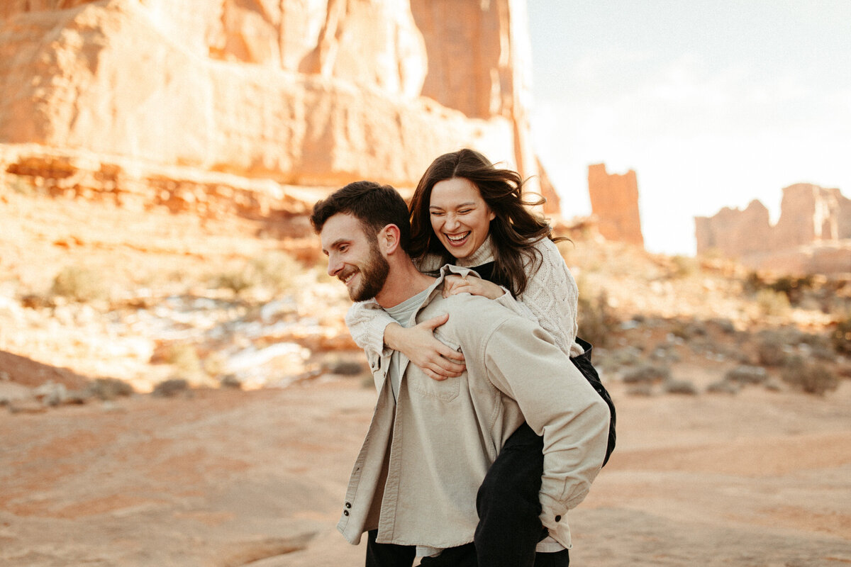 A girl is riding piggy back on her fiancé's back with the desert behind them,
