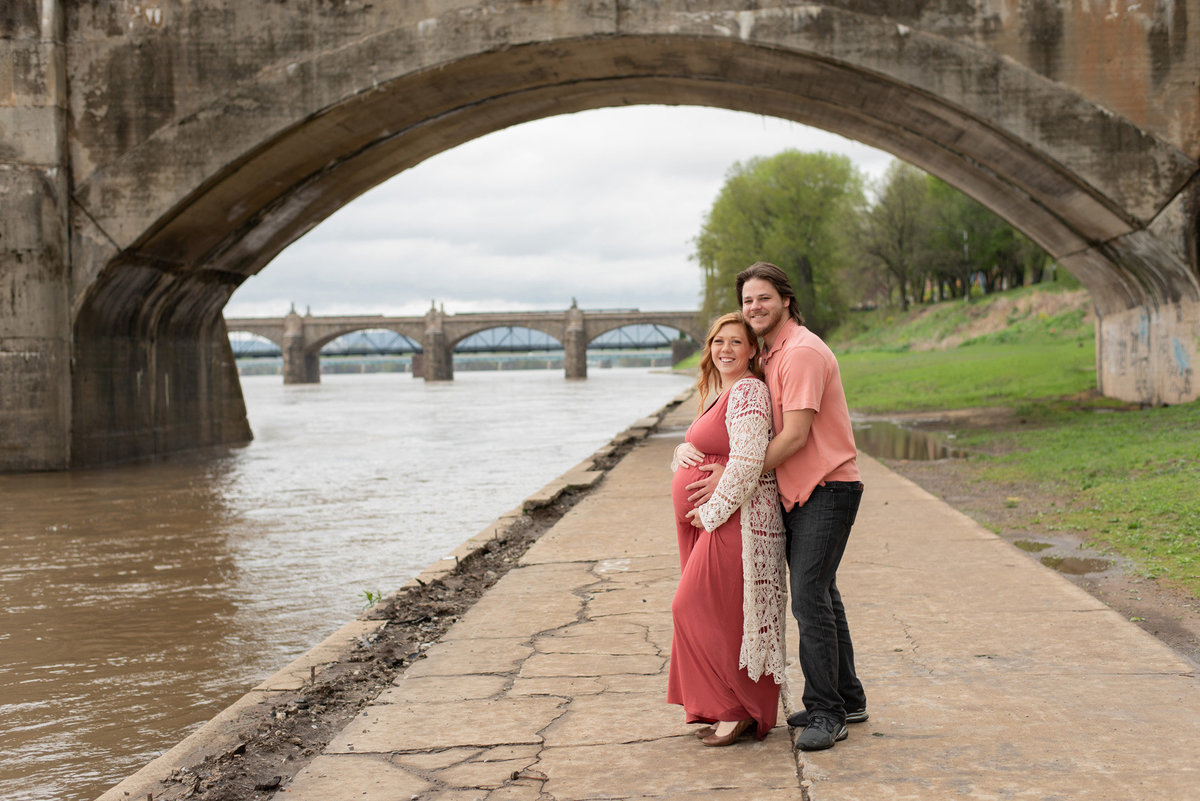 Husband & Pregnant Wife  posed along river with bridges behind them