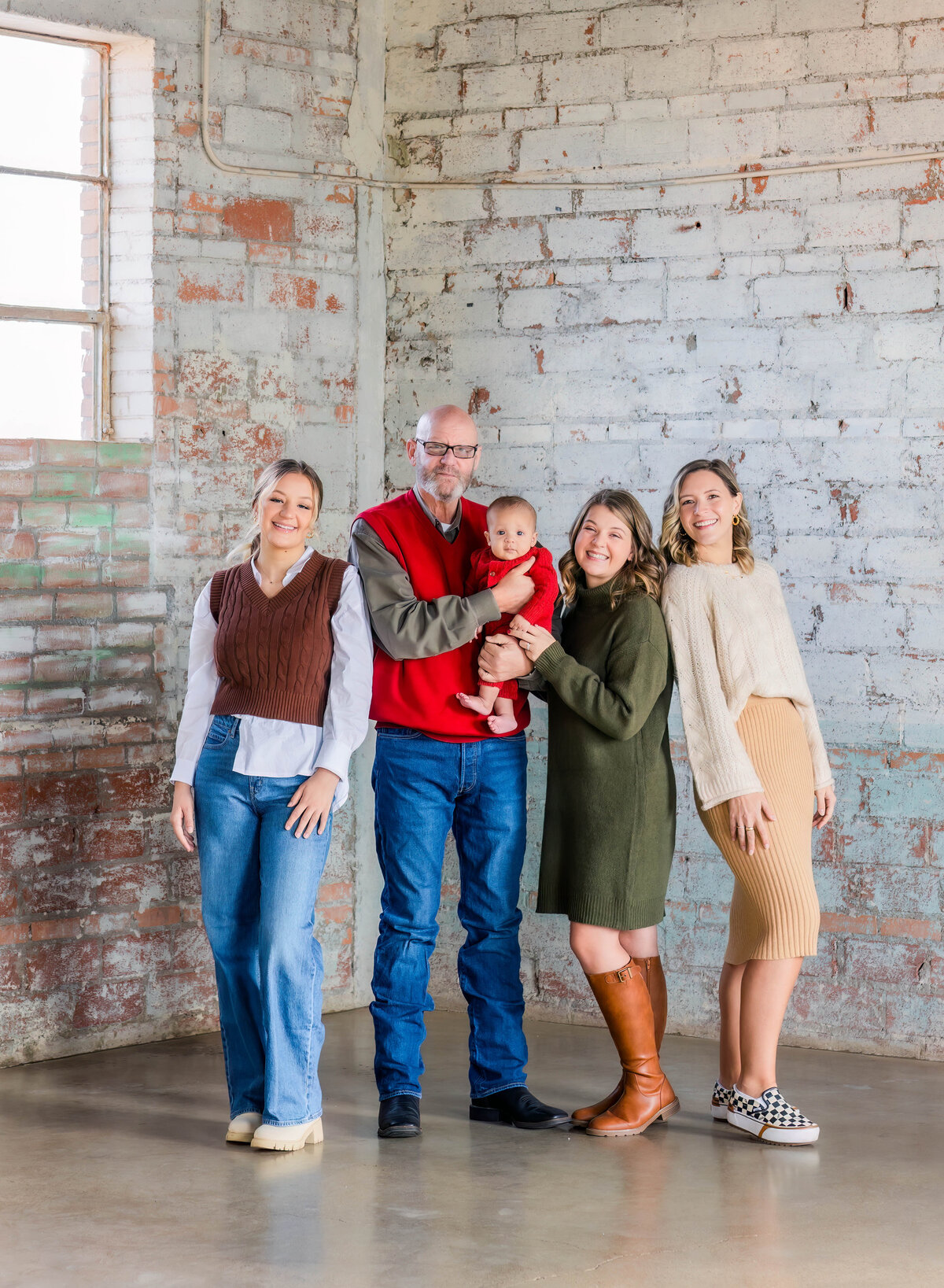 Dad posed with 3 daughter's and grandson with all looking at the camera during an indoor winter family session.  One  woman is wearing a brown vest over a white long sleeved blouse with jeans, man is wearing a red vest over an olive shirt with jeans.  another woman is wearing an olive green sweater dress with brown leather boots and the other woman is wearing a cream sweater with a khaki skirt and checkered vans.  The baby is wearing a red sweater romper.