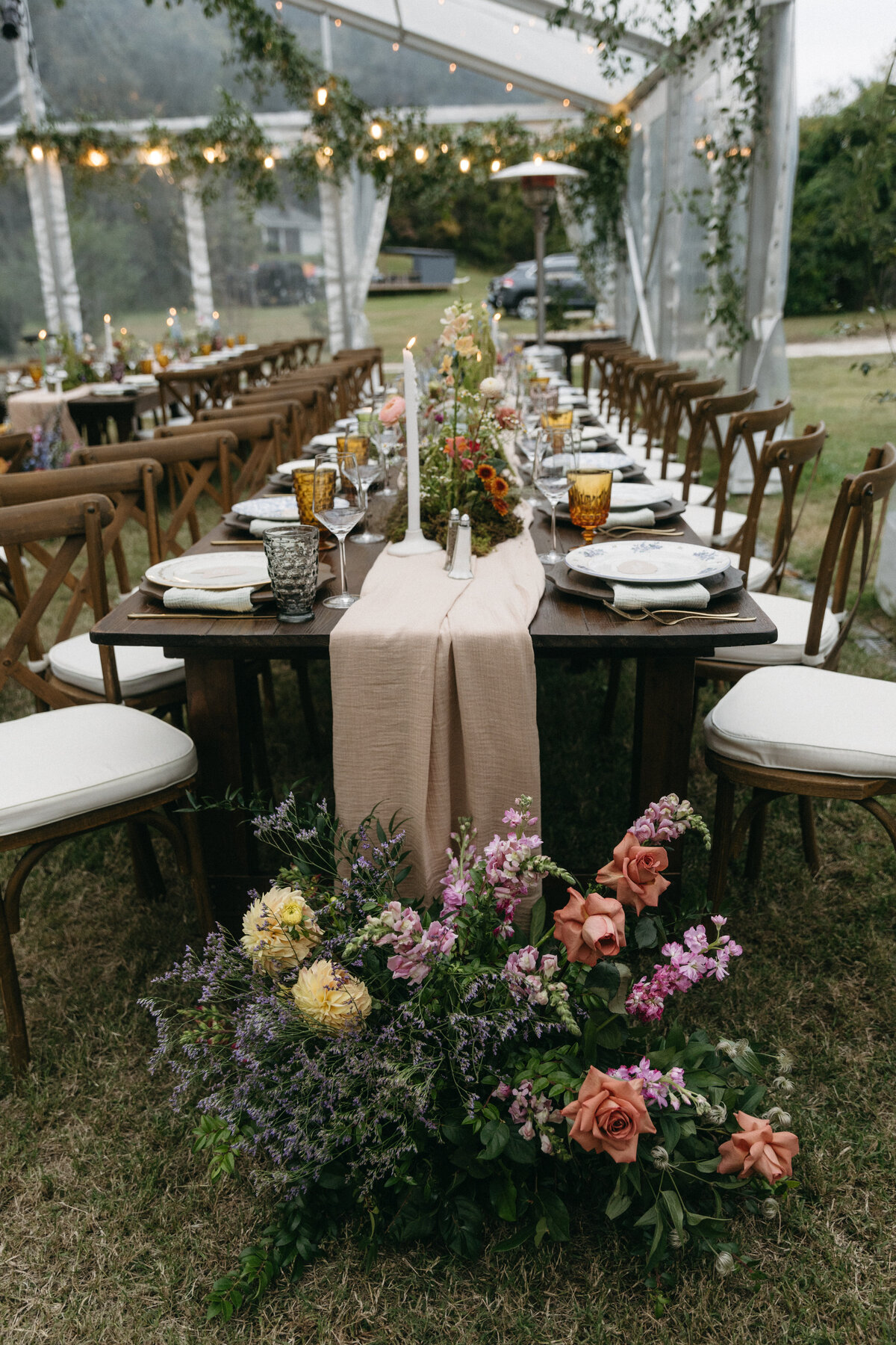Long outdoor wedding reception table under a clear tent adorned with lush garden-style florals, pastel taper candles, amber glassware, and a soft blush table runner, designed by a Northwest Arkansas wedding florist to create an elevated woodland-meets-storybook aesthetic with abundant seasonal flowers at the table’s base.