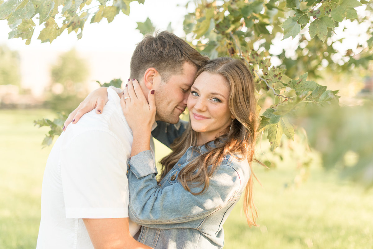 Engaged couple hugging as girl smiles at camera and guy rests his forehead against the side of her face underneath a tree.