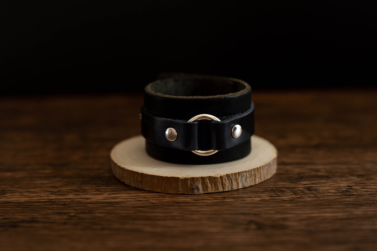 A black leather bracelet with metal studs, displayed on a wooden stand against a dark background.
