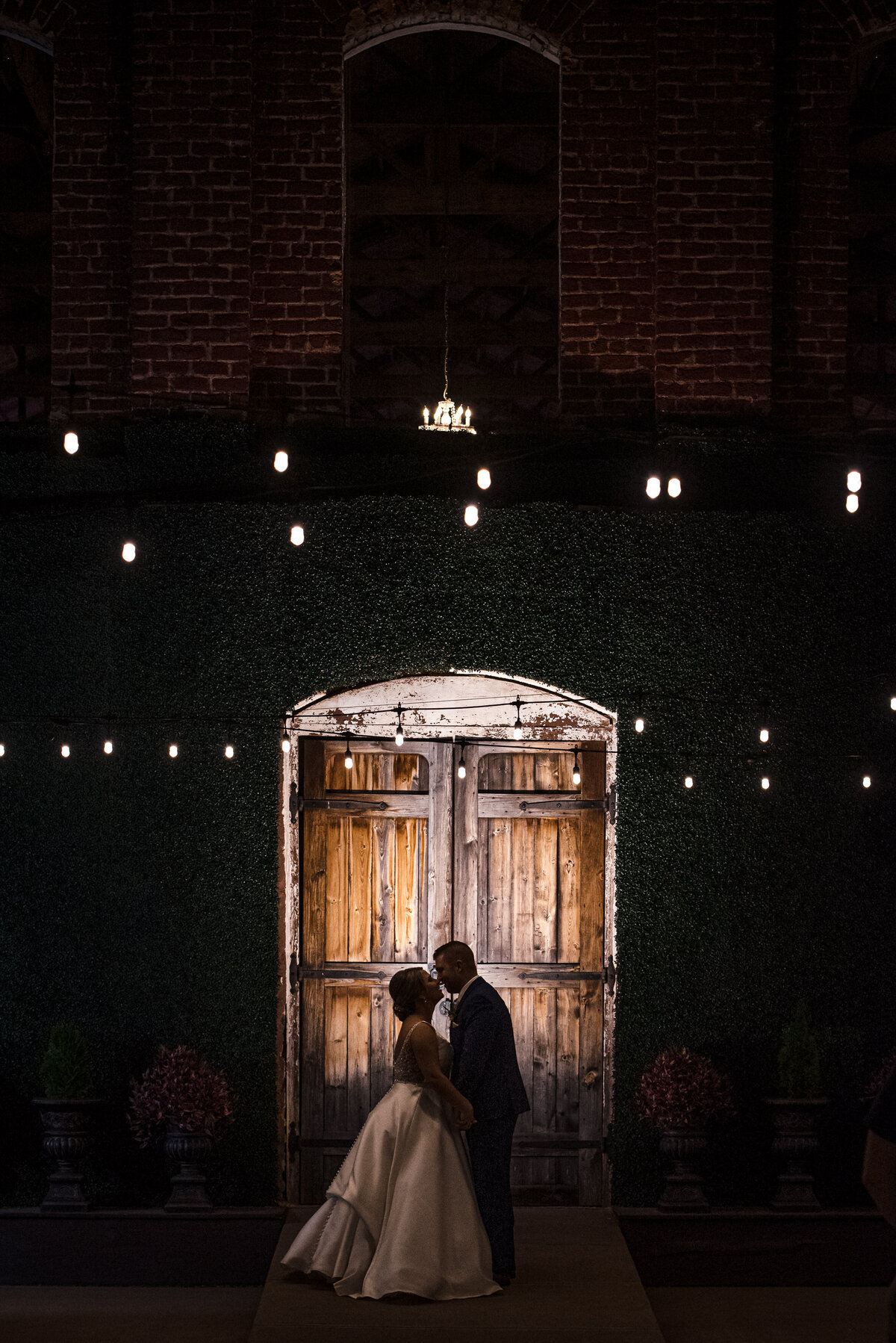 Silhouette-of-bride-and-groom-at-night-in-front-of-the-wooden-doors-under-the-fairy-lights-at-The-Providence-Cotton-Mill